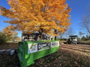 A green Dump-It Dumpster Rentals dumpster placed in a residential yard with a vibrant orange autumn tree in Concord, NH.