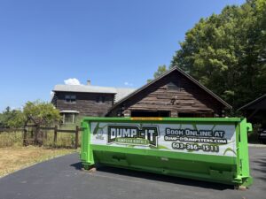 A green Dump-It Dumpster Rentals dumpster placed in a driveway in front of a rustic wooden house in Concord, NH.