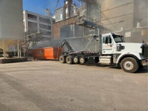 A Rushmore Dumpster truck servicing an orange roll-off dumpster at an industrial site in Rapid City, SD.
