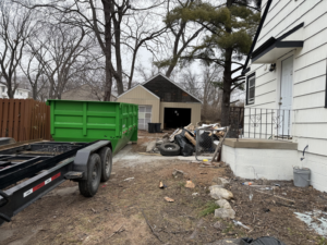 A green dumpster trailer placed next to a house with a large pile of construction and yard debris for Proper Dumpster Rentals in Kansas City, KS.