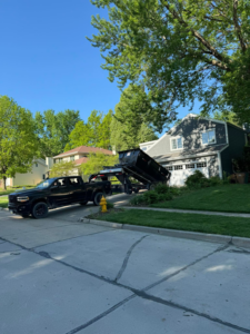 A black pickup truck delivering a dumpster trailer to a residential driveway for Waste Warriors Dumpster Rental of Des Moines, IA.