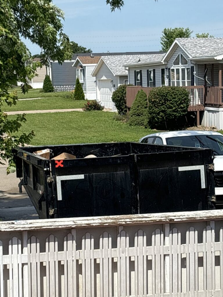 A black dumpster trailer partially filled with junk in a residential neighborhood, provided by Waste Warriors Dumpster Rental of Des Moines, IA.