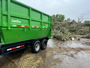 A green dumpster trailer positioned next to a large pile of tree branches and logs for debris removal by Proper Dumpster Rentals in Kansas City, KS.