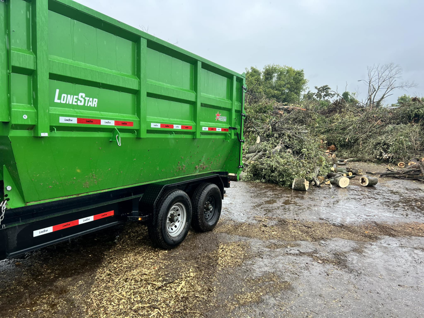 A green dumpster trailer positioned next to a large pile of tree branches and logs for debris removal by Proper Dumpster Rentals in Kansas City, KS.