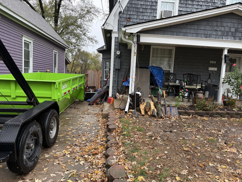 A green dumpster trailer parked next to a residential house with a pile of junk and debris for Proper Dumpster Rentals in Kansas City, KS.