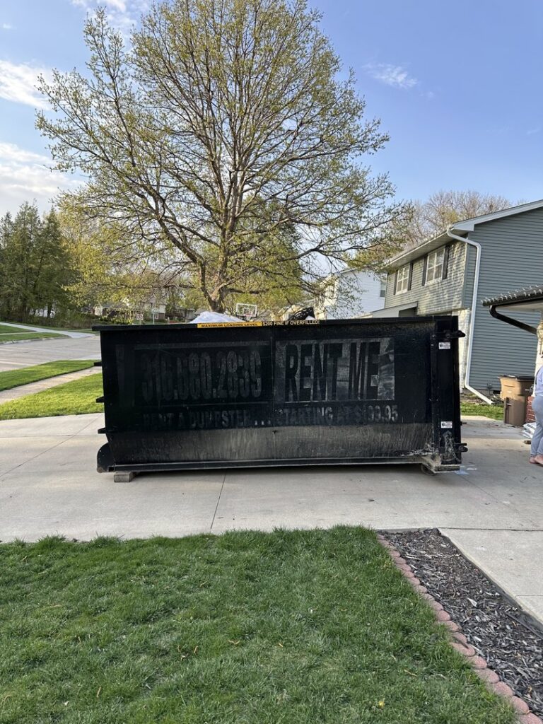 A black dumpster trailer with "RENT-ME" on its side, parked in a residential driveway by Waste Warriors Dumpster Rental of Des Moines, IA.