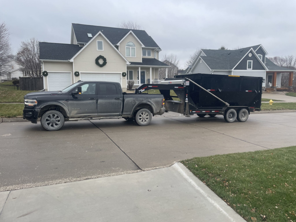 A black pickup truck picking up a full dumpster trailer from a residential home for Waste Warriors Dumpster Rental of Des Moines, IA.