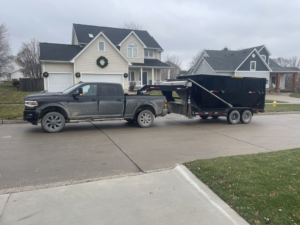 A black pickup truck picking up a full dumpster trailer from a residential home for Waste Warriors Dumpster Rental of Des Moines, IA.