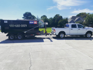 A white pickup truck towing a black dumpster on a trailer down a residential street for The Dumpster Dad in Wilmington, NC.