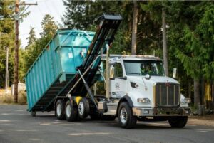 A white truck loading a teal roll-off dumpster, demonstrating junk removal services in Sterling Heights, MI.