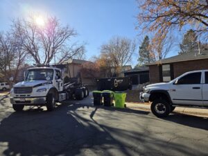 A Humpty Dump Roll-Offs & Dumpsters truck with a dumpster on a residential street, with trash bins nearby in Commerce City, CO.