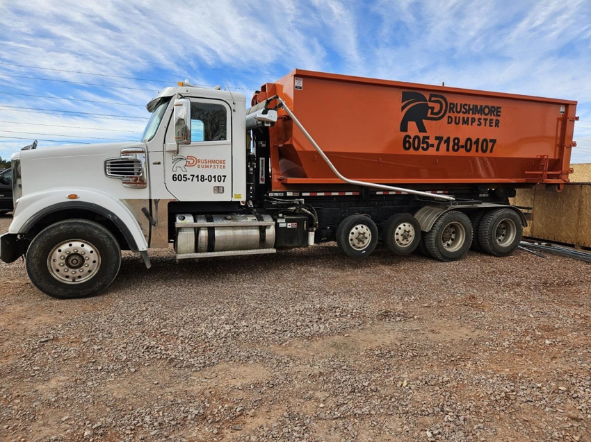 A white Rushmore Dumpster truck with an orange roll-off dumpster attached, parked on a gravel lot in Rapid City, SD.