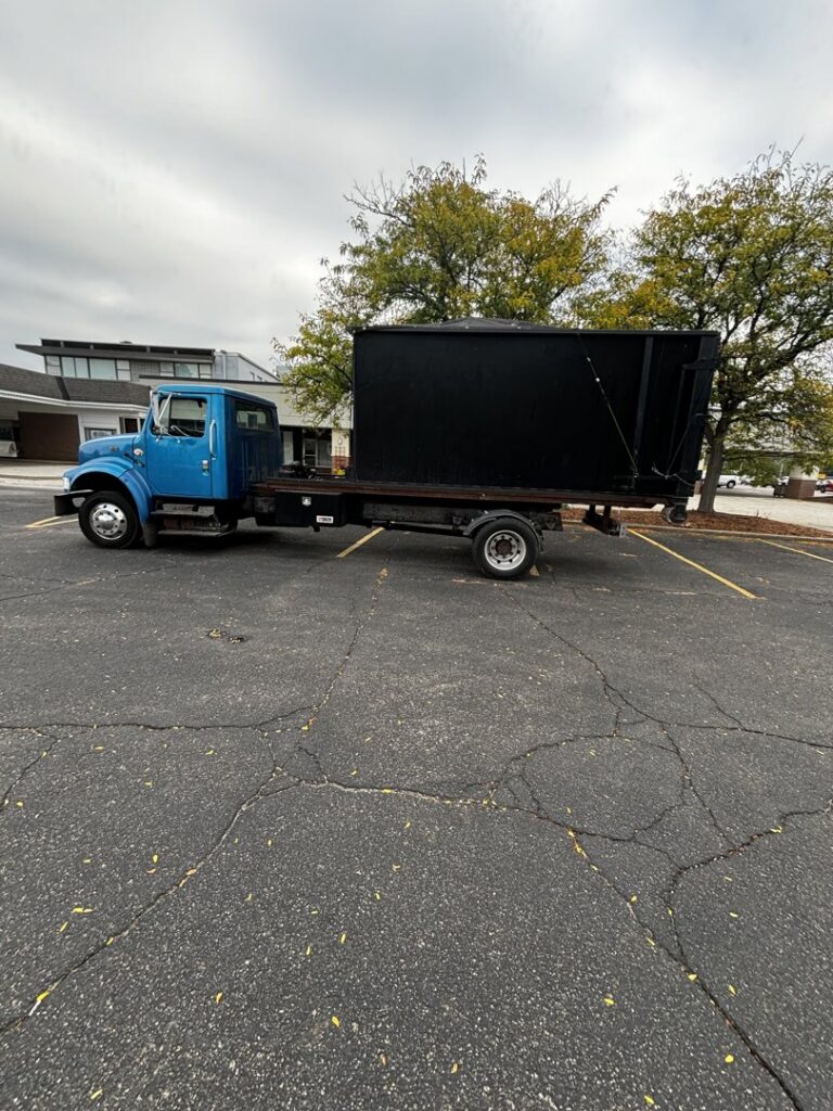 A blue roll-off truck with a black dumpster on its bed, parked in a commercial parking lot by Wagners Property Services LLC in Canton, OH.