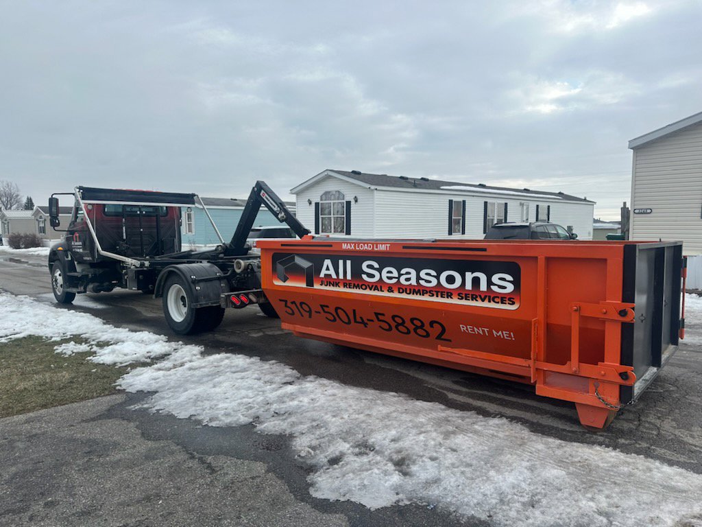 An orange dumpster attached to a truck driving on a snowy road, provided by All Seasons Junk Removal & Dumpster Services in Center Point, IA.