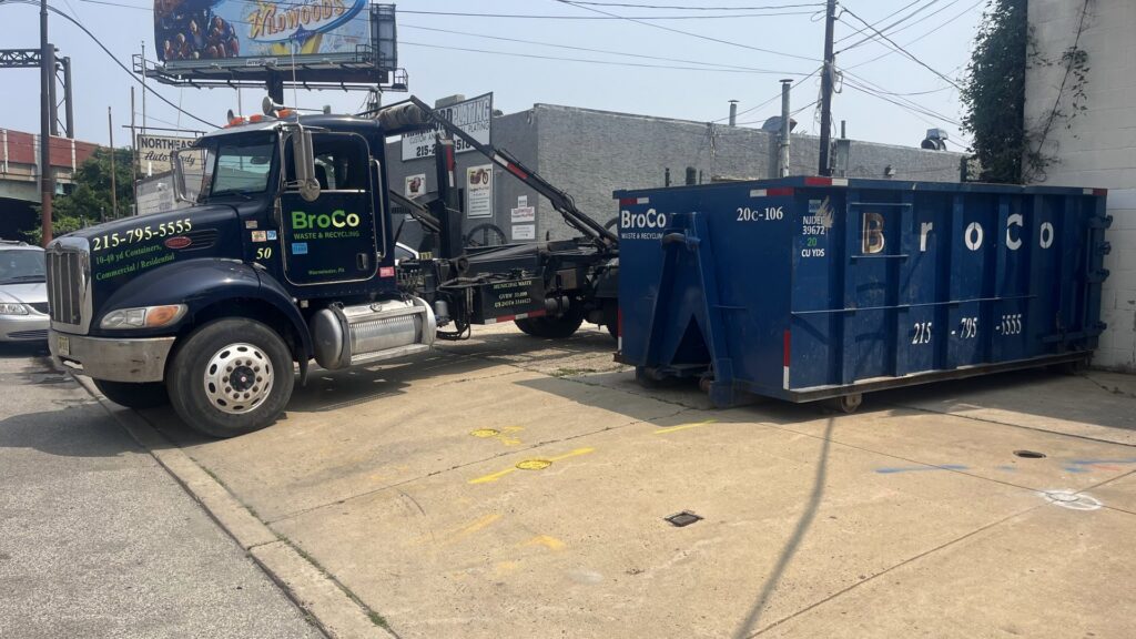 A Broco Waste & Recycling truck unloading a blue dumpster at a commercial site in Warminster, PA.