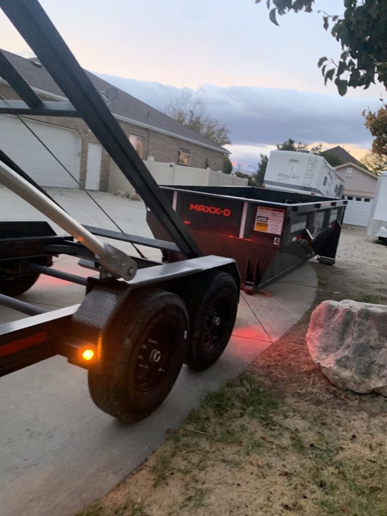A RAAAM Dumpster Rentals roll-off dumpster being unloaded at dusk in a residential area of Salt Lake City, UT.