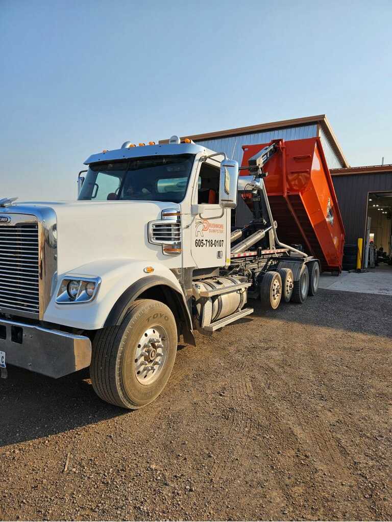 A Rushmore Dumpster truck unloading an orange roll-off dumpster at a waste facility in Rapid City, SD.