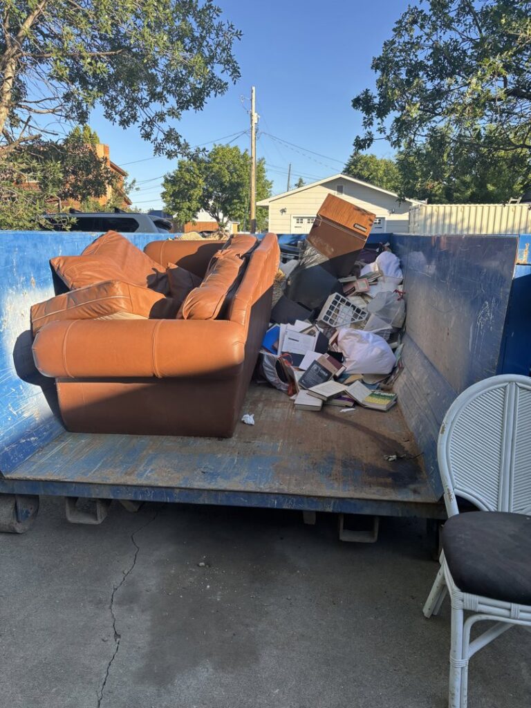 A blue roll-off dumpster containing a couch and various junk items for removal by Evergreen Disposal in Kalispell, MT.