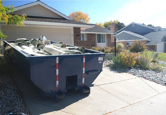 A blue roll-off dumpster filled with rolled-up carpets and other debris in a residential driveway for junk removal in Sterling Heights, MI.