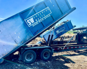 Two blue dumpsters from Superior Waste Management being loaded or unloaded from a trailer for junk removal services in Amory, MS.