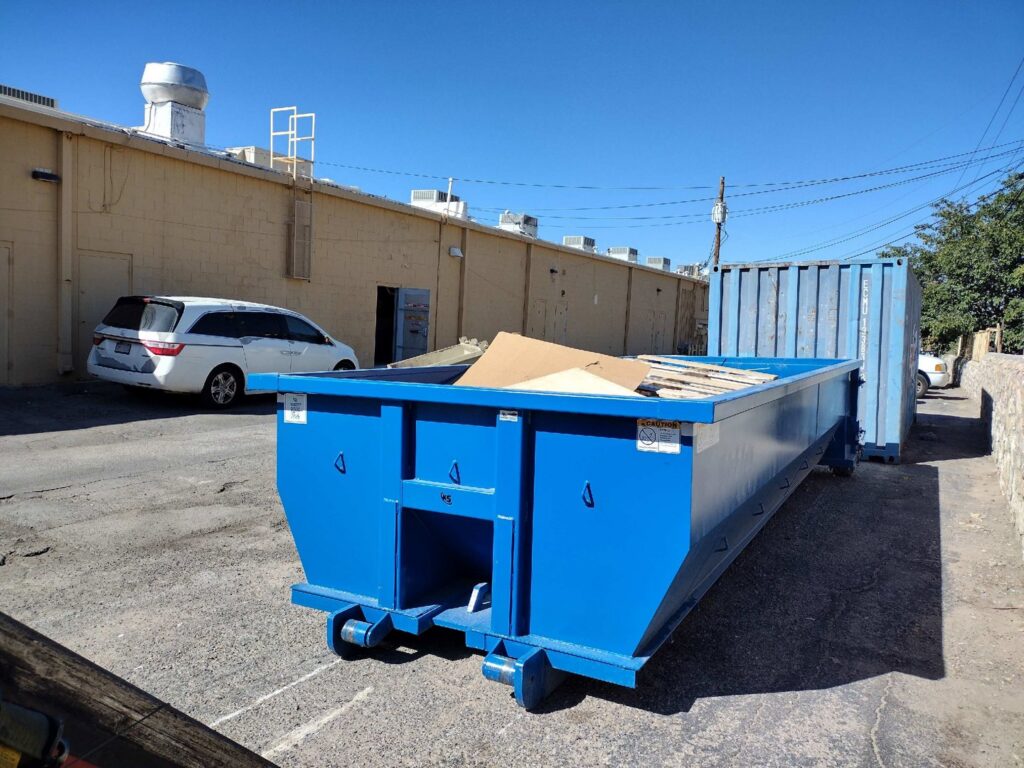 Two blue dumpsters, one partially filled with cardboard and debris, at a commercial site for Tidy up time junk removal in El Paso, TX.