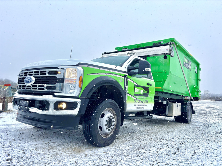 A DumpStor of Louisville truck with a green roll-off dumpster driving on a snowy road for junk removal service in Louisville, KY.