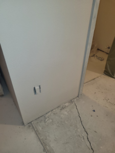 A wall with a new electrical outlet cutout and cracked floor, showing handyman work by Precision Craftsman LLC in San Antonio, TX.