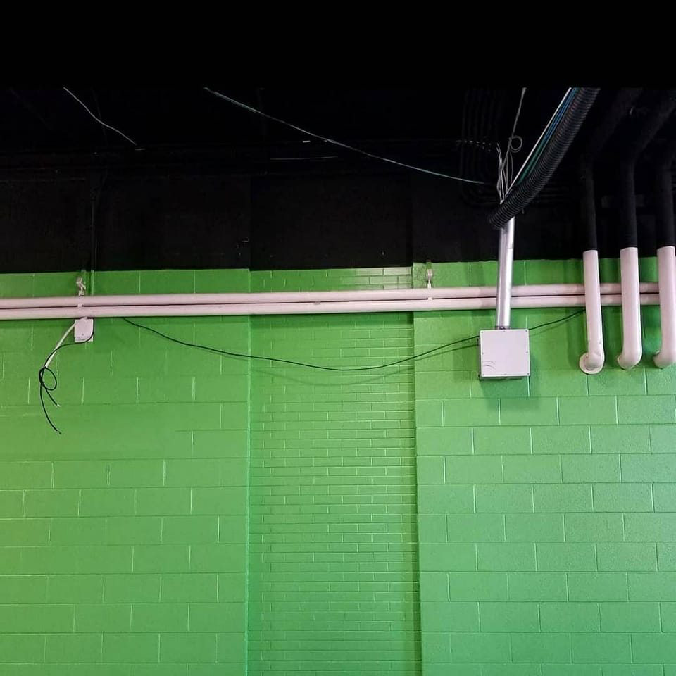 Exposed electrical wiring and conduit with pipes on a green brick wall, showing handyman electrical and plumbing work by Reshaud Renovation LLC in Richmond, VA.