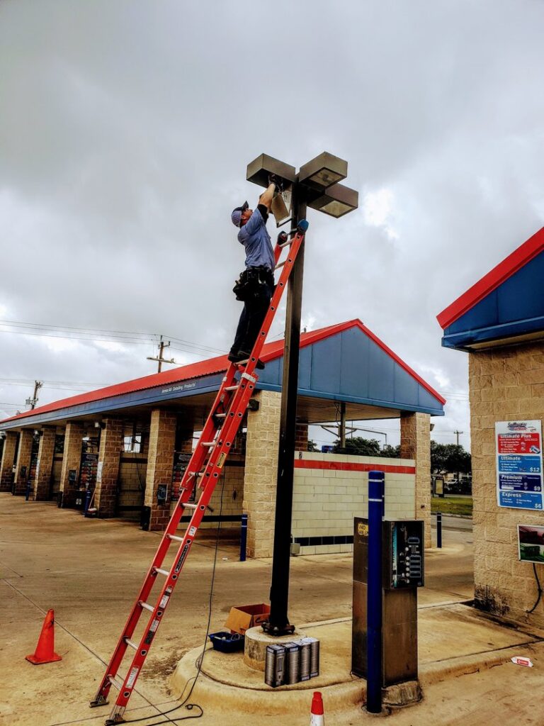 An electrician on a ladder repairing an outdoor light fixture at a commercial property for Denali Electric LLC in San Antonio, TX