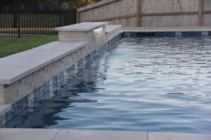 An elegant pool waterfall feature with stone coping, designed by Limitless Custom Pools and Backyards in Houston, TX.