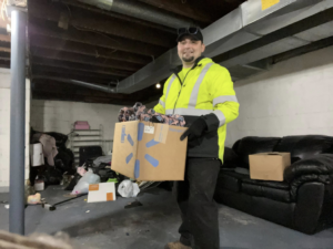 An employee from 2 Much Junk? Junk Removal carrying a box of items from a cluttered basement in Grand Rapids, MI.
