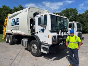 A One Waste Solutions employee in a hard hat and safety vest standing proudly in front of two garbage trucks in Murfreesboro, TN.