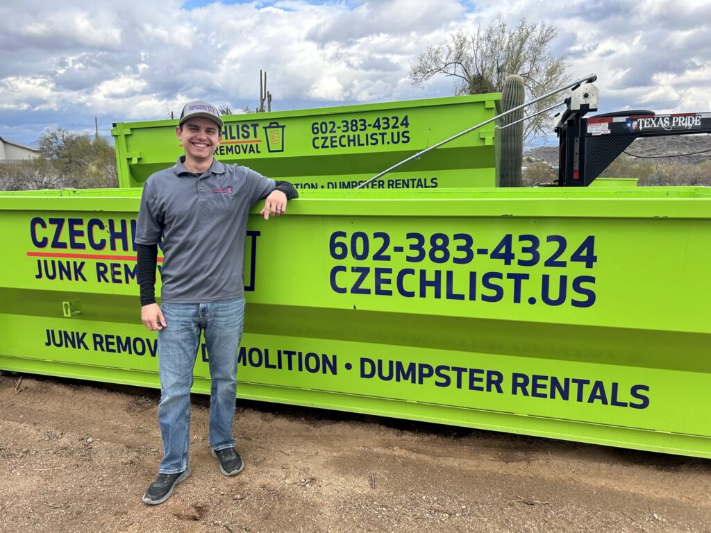 An employee standing proudly next to two bright green junk removal dumpsters from CzechList Junk Removal in Scottsdale, AZ.