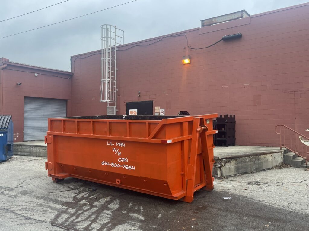 An empty orange dumpster with the business name visible, placed next to a commercial building by Lil Man W/A Can LLC in Columbus, OH.