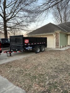An empty black dump trailer from D's Dumpster Rentals parked in a residential driveway in Overland Park, KS.