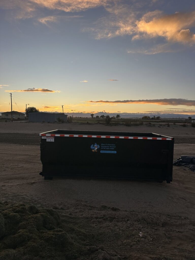 An empty dumpster from Blue J's Services Dumpster Rental placed in a field at sunset in Greeley, CO.