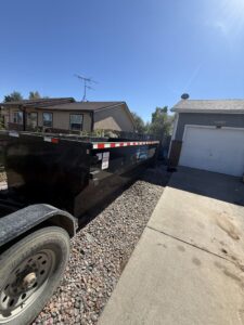 An empty dumpster from Blue J's Services Dumpster Rental placed on a gravel area next to a residential garage in Greeley, CO.