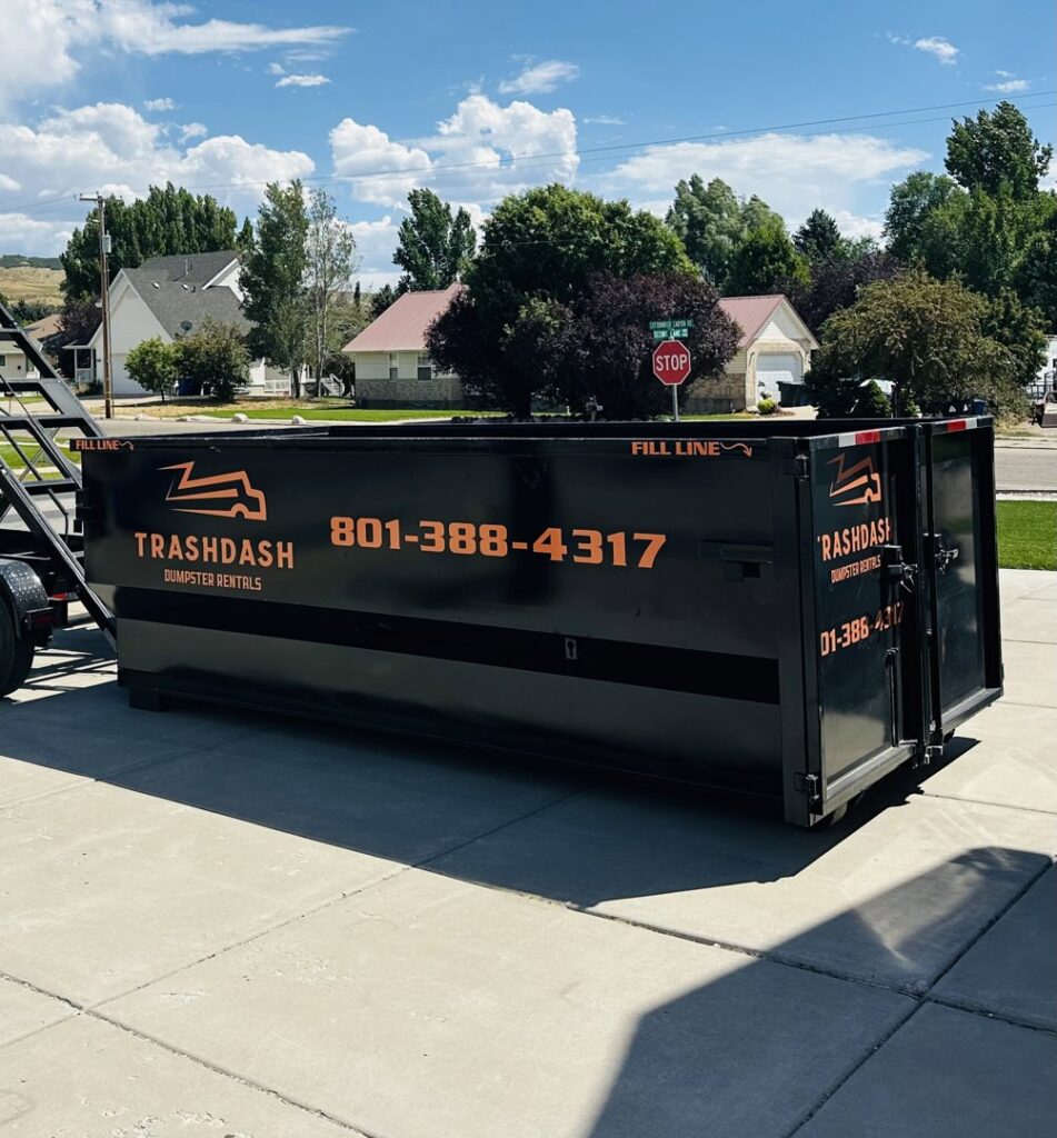 An empty TrashDash dumpster on a trailer parked in a residential driveway in West Haven, UT.
