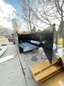 An empty junk removal dumpster with its back open, next to rolled carpet and debris, provided by Pack N Haul in West Valley City, UT.