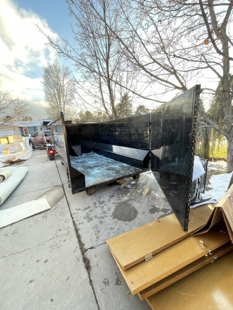 An empty junk removal dumpster with its back open, next to rolled carpet and debris, provided by Pack N Haul in West Valley City, UT.