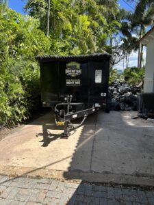 An empty Driveway Safe Dumpster Rental trailer parked next to a pile of junk, ready for removal in Fort Lauderdale, FL.