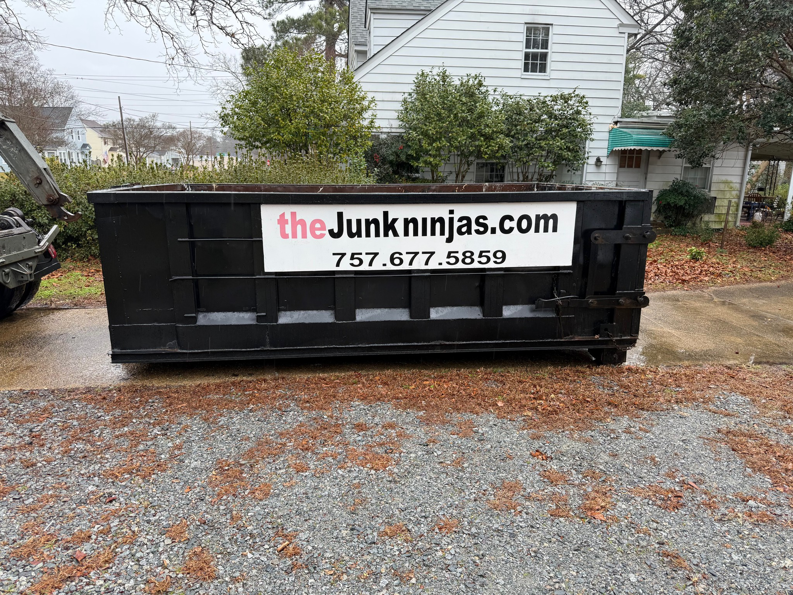 An empty black dumpster from The Junk Ninjas being delivered or picked up on a gravel driveway in Hampton Roads, VA.