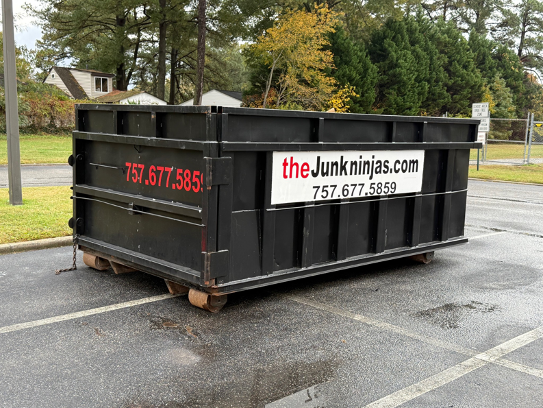 An empty black dumpster from The Junk Ninjas parked in a commercial parking lot in Hampton Roads, VA.