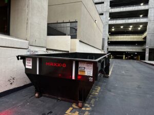 An empty RAAAM Dumpster Rentals dumpster placed in an urban setting next to a parking garage in Salt Lake City, UT.