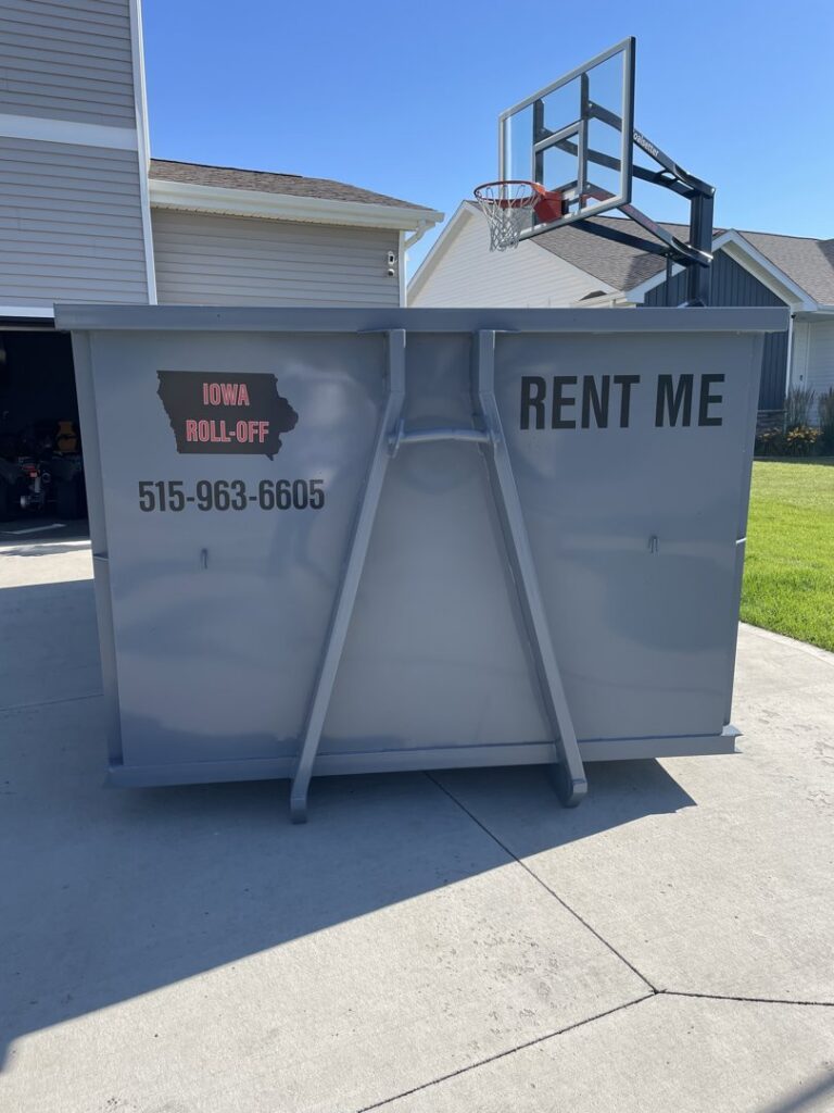 An empty grey roll-off dumpster from Iowa Roll Off placed in a residential driveway with a basketball hoop in the background in Ankeny, IA.