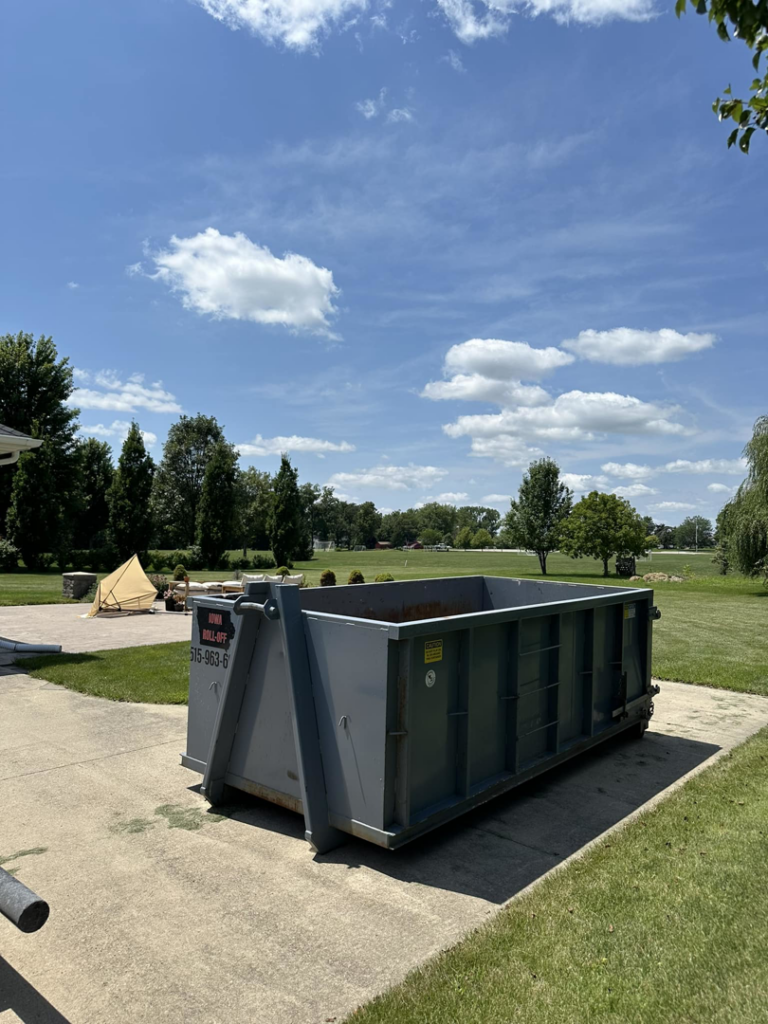 An empty grey roll-off dumpster from Iowa Roll Off placed on a concrete pad in a residential backyard in Ankeny, IA.