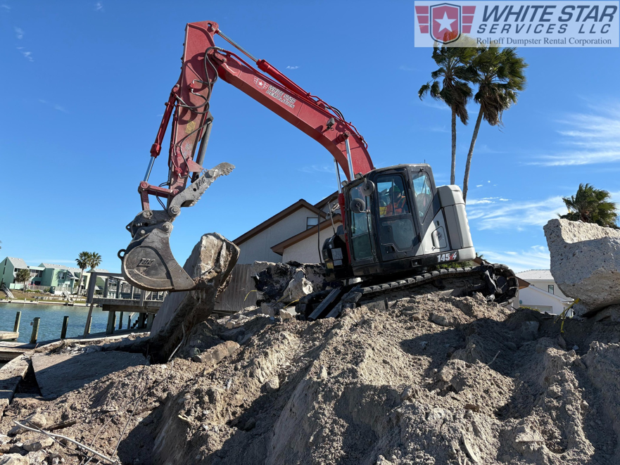 An excavator from White Star Services LLC clearing concrete debris next to a waterway in Corpus Christi, TX.