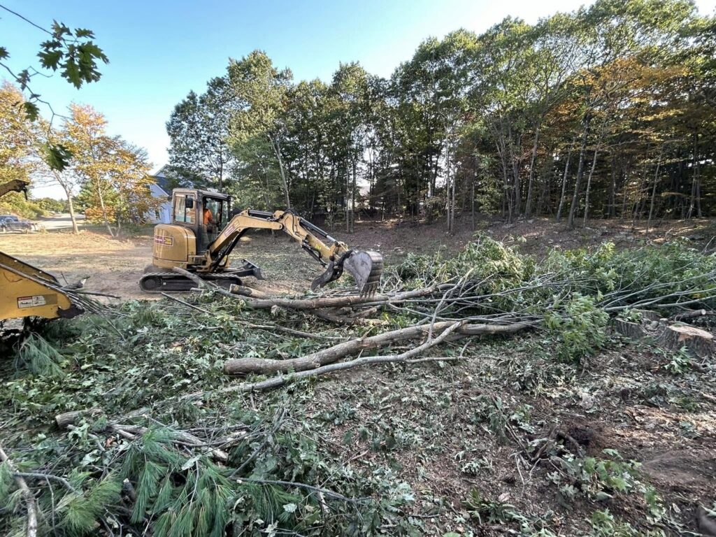 An excavator clearing tree debris and branches during land clearing by JTE & Company in Oxford, MA.