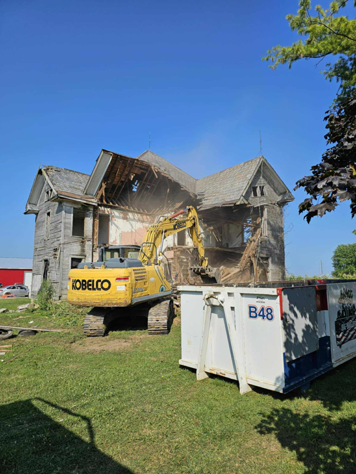 An excavator demolishing an old house with a Bragg About This Dump roll-off dumpster nearby in Columbus, OH.