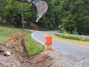An excavator bucket digging a trench alongside a road, indicating excavation services by Huntington Home Repair in Huntington, WV.